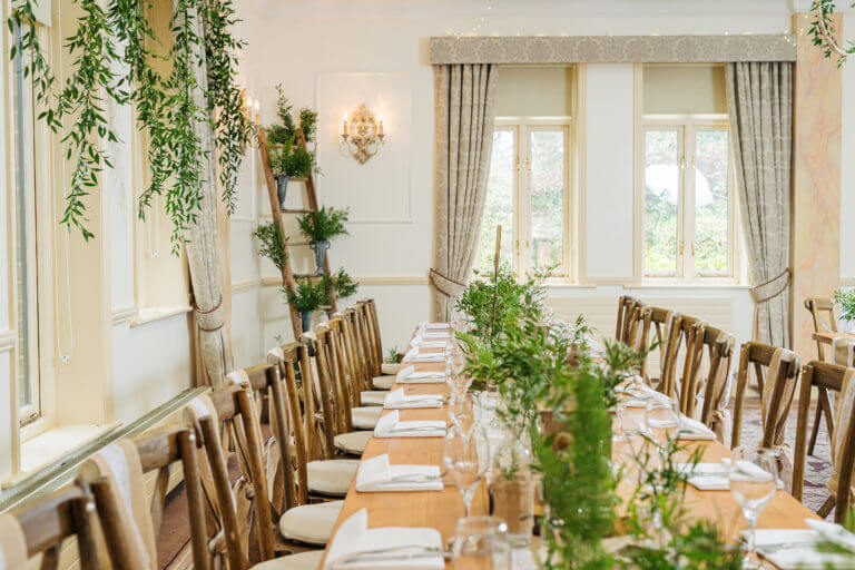 A room is set up for a wedding breakfast with long tables and lots of green foliage used as decor