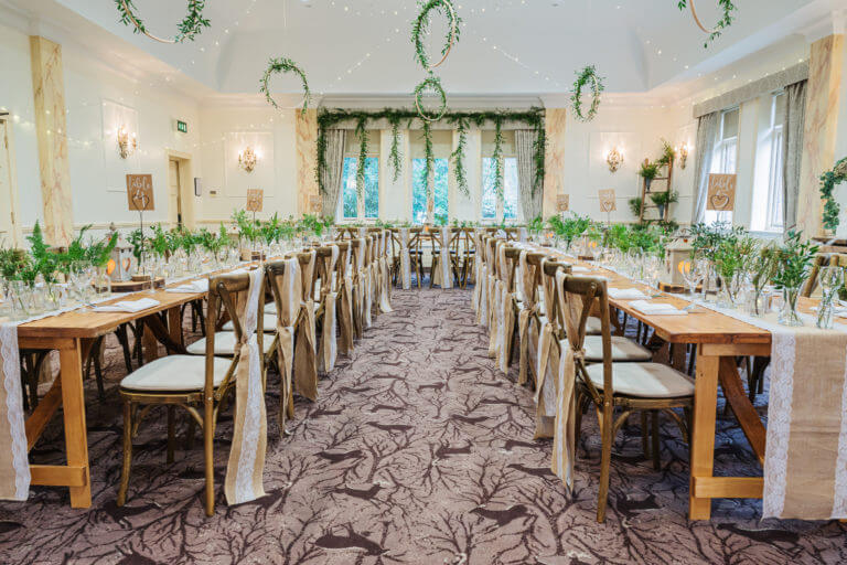 A room is set up for a wedding breakfast with long tables and lots of green foliage used as decor