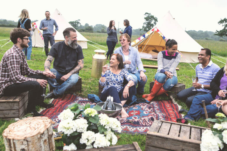 Group of colleagues in casual clothing enjoying glasses of Prosecco and chatting in a field with tents