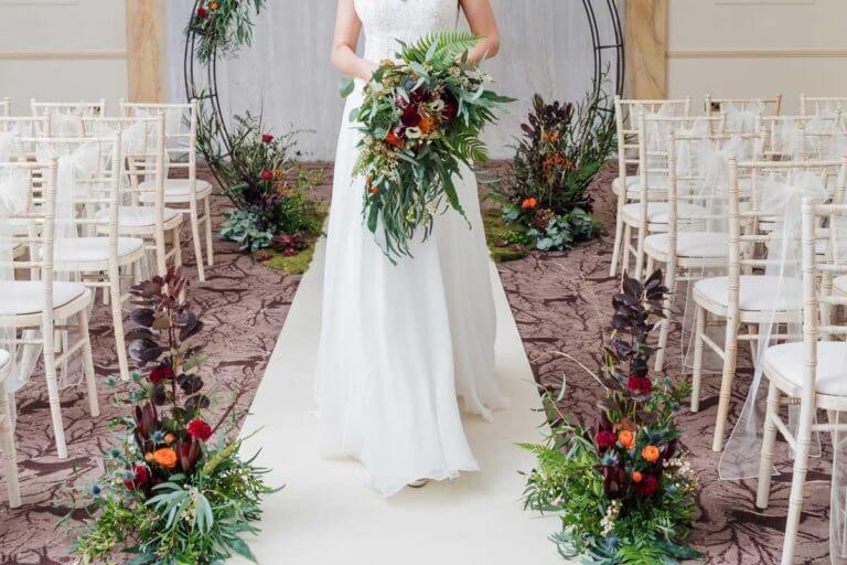Bride walks up the white carpet aisle holding big bouquet with red flowers
