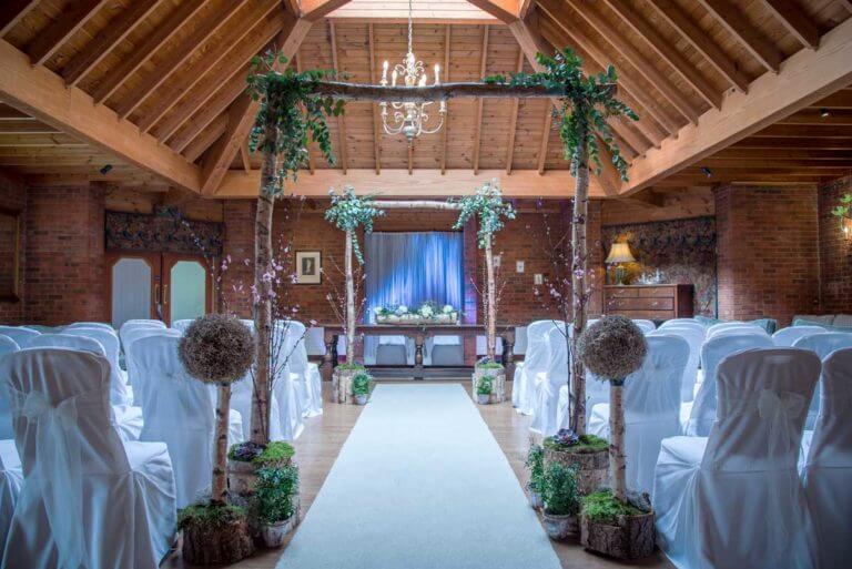 A room with wood panelled ceiling set up for a wedding ceremony with a white aisle runner, white banqueting chairs and two woodland themed arches