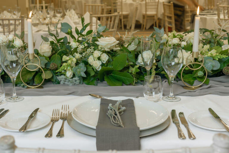 A tablescape with white plates, grey napkins and silver currently. Running down the center of the table is a low flower arrangement with white roses and foliage.