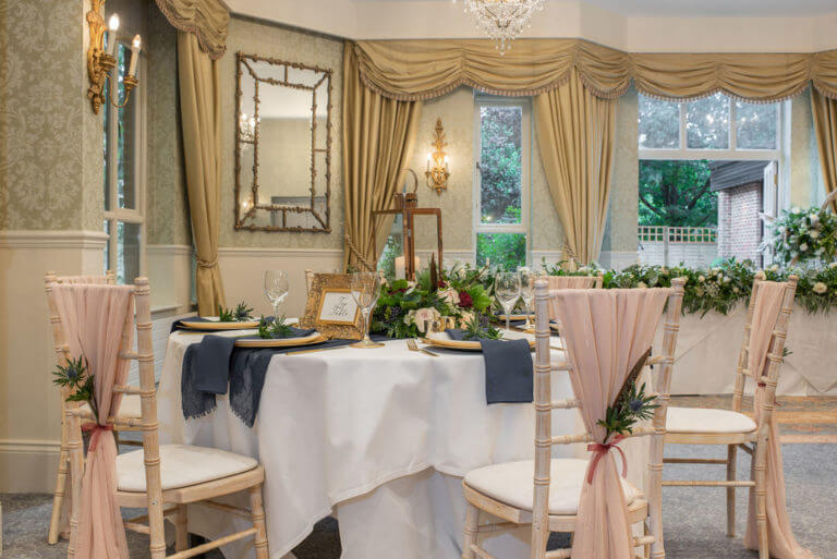 Room set up for wedding breakfast with floral arrangement and pink sashes on chairs