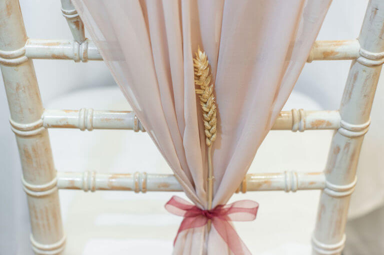 Detail of a blush-pink wedding sash on a chair with a dried sprig of barley tied with a dark pink ribbon.