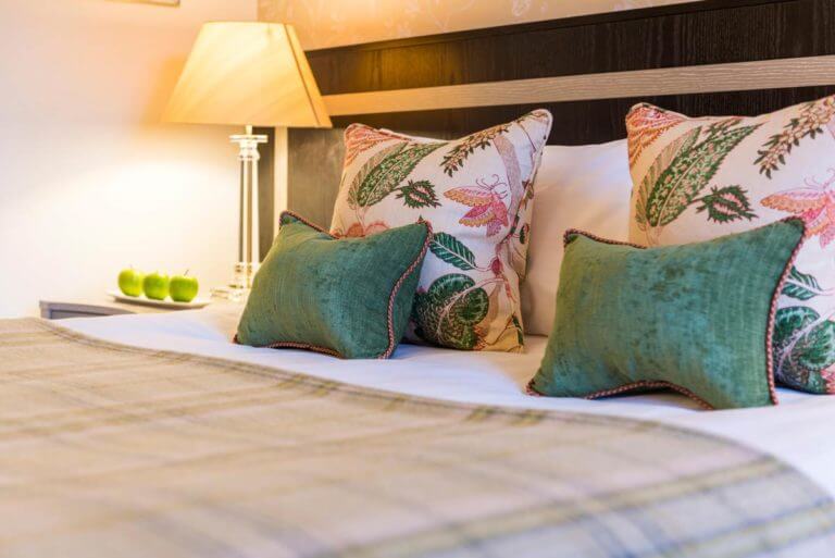 Bed in hotel room with two small green cushions and two large nature patterned cushions in green and pink. Side table with lamp and green apples