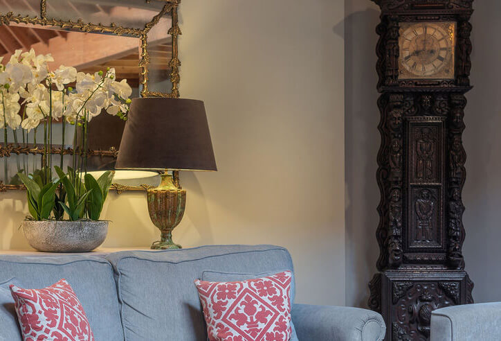 Detail shot of a lounge area with a grey sofa with red patterned cushions, a large grandfather clock, side table with white flowers and lamp and large mirror with gold frame detail