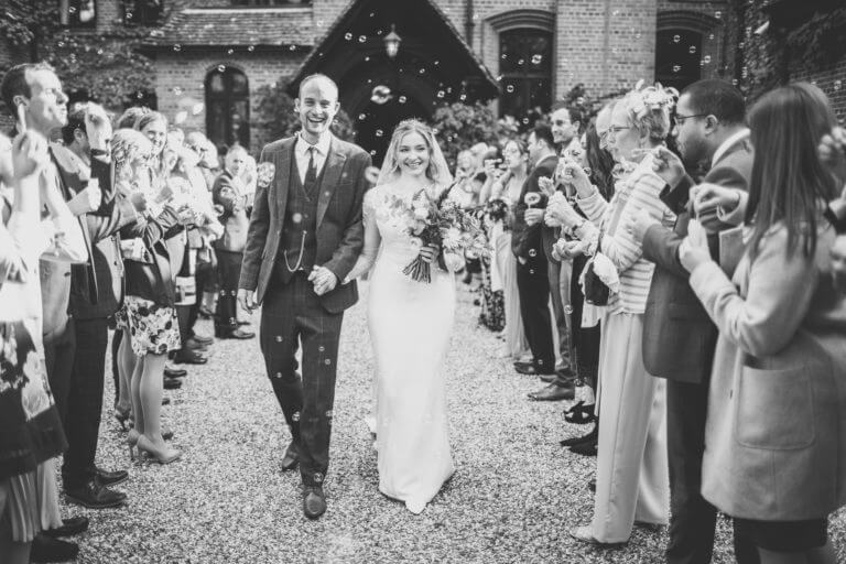 Black and white photo of bride and groom leaving wedding venue surrounded by happy guests blowing bubbles