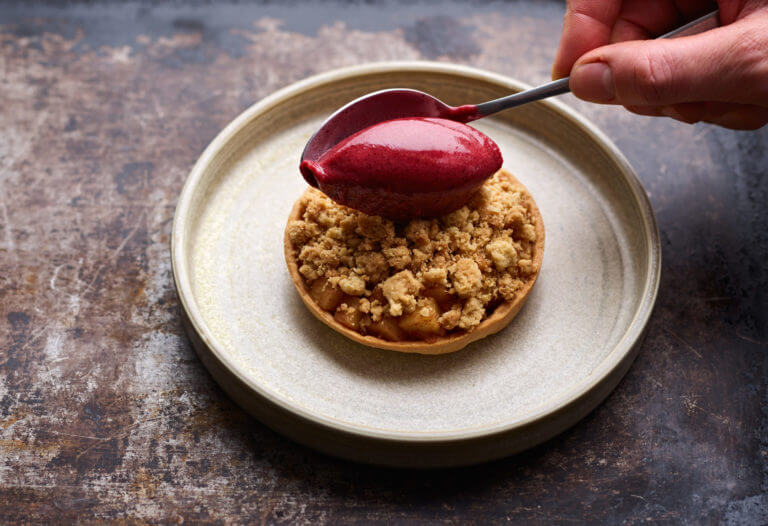Apple crumble tart on a plate with hand scooping berry sorbet on top
