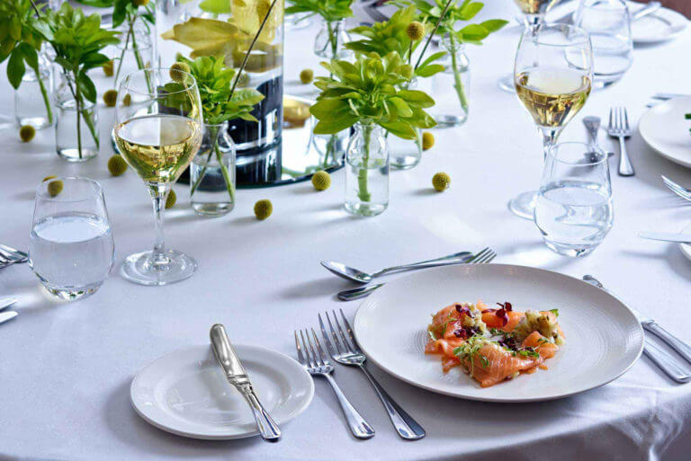 Table set with white cloth for meal with two glasses of white wine and a smoked salmon dish, lots of foliage in small bottles as centrepiece