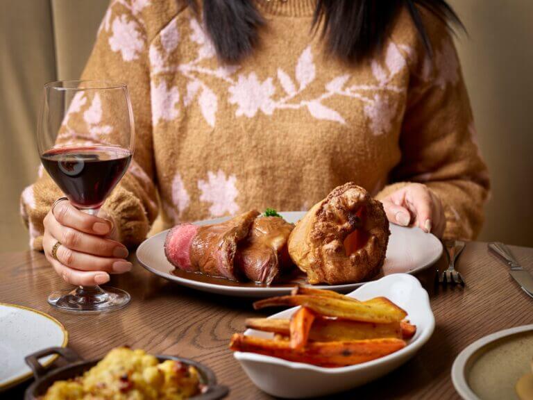 Woman holds glass of red wine and prepares to tuck into beef Sunday roast on wooden table in Cambium restaurant, Brockenhurst