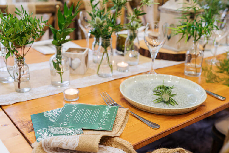 A wedding table setting with a blue ceramic plate with a thistle placed on it and green wedding invitations to the side