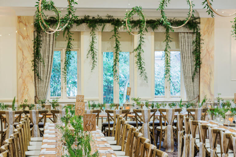 A light filled room set up with long wooden trestle tables filled with folliage table centers for a wedding. Wooden hoops filled with foliage hang from the ceiling.