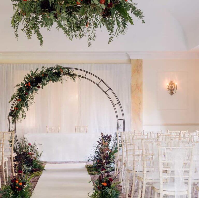A room set up for a wedding ceremony with an autumn flower filled moon arch, white carpet runner and chairs with white sashes.