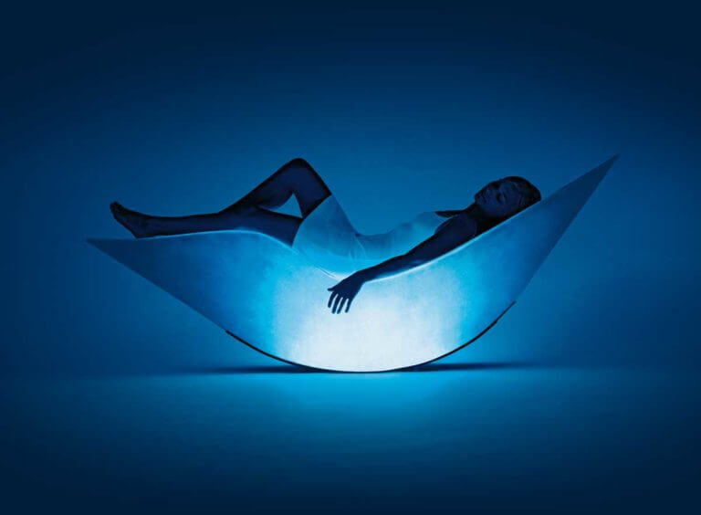 Woman wearing white slip dress lies on Alphasphere Deluxe bed enjoying relaxing immersive experience with blue lights