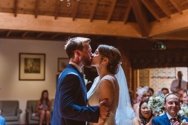 Bride and groom kiss during wedding ceremony with happy wedding guests looking on in lounge room with wood panelled ceiling