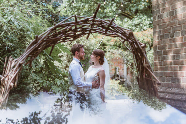 Bride and groom stand under rustic archway made of sticks in hotel wedding venue grounds, looking lovingly at each other