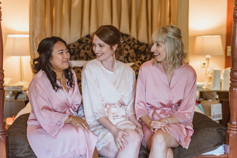 Bride sitting on bed in white silk robe with two bridesmaids wearing pink silk robes