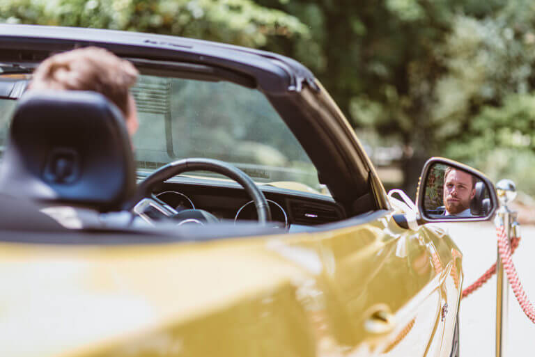 Groom drives yellow car on wedding day with face shown in wing mirror reflection