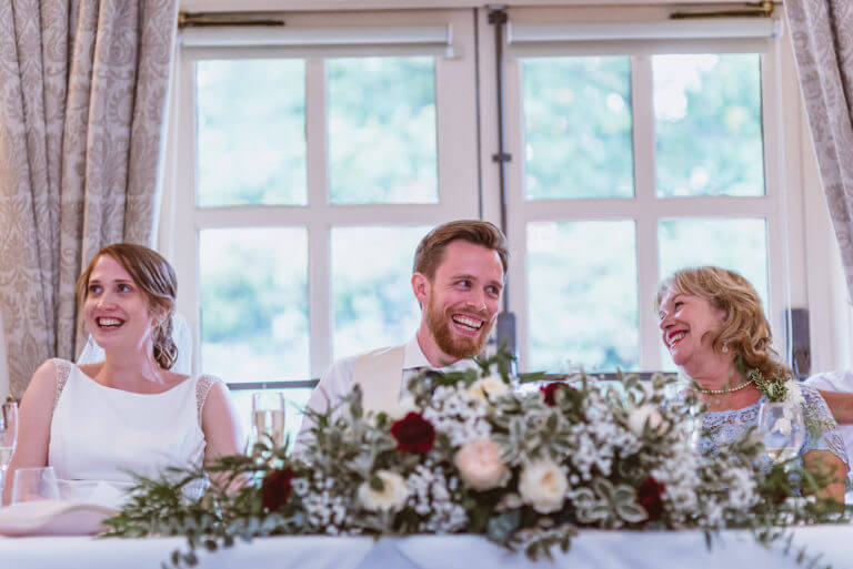 Bride, groom and mother sit on top table laughing during wedding, with red and white flowers and foliage on table