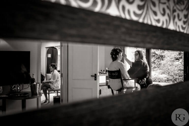 Black and white photo of bride getting makeup professionally done through reflection of a mirror
