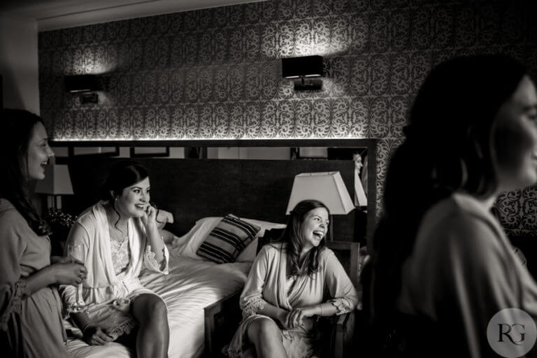 Black and white photo of bride sitting on bed in hotel room with bridal party having a drink and laugh