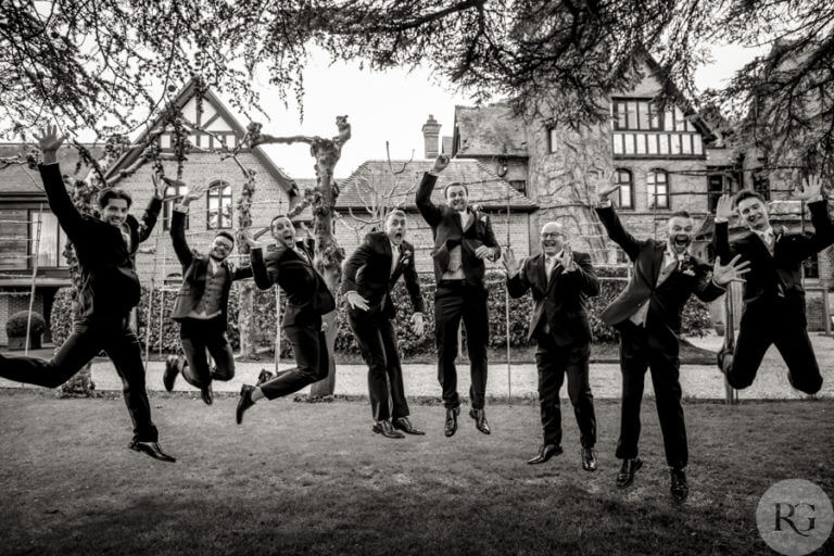 Black and white action shot of groom and groomsmen jumping in the air on the lawn outside hotel wedding venue Careys Manor Hotel & SenSpa