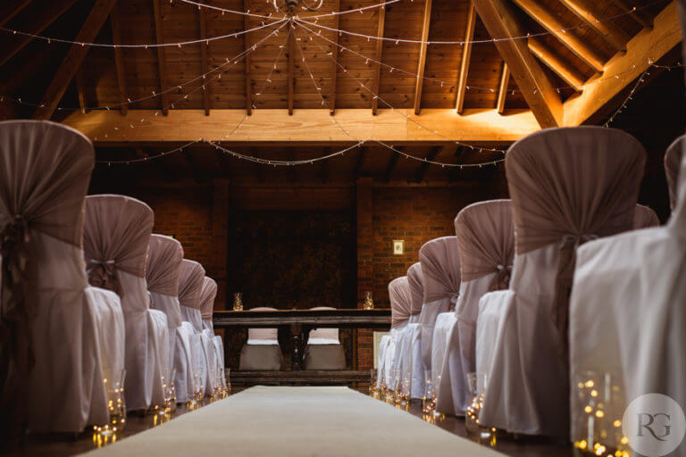 Aisle view of hotel lounge set for wedding ceremony in room with wood panelled ceiling, white banqueting chairs with brown sashes and fairy lights in jars lining the aisle