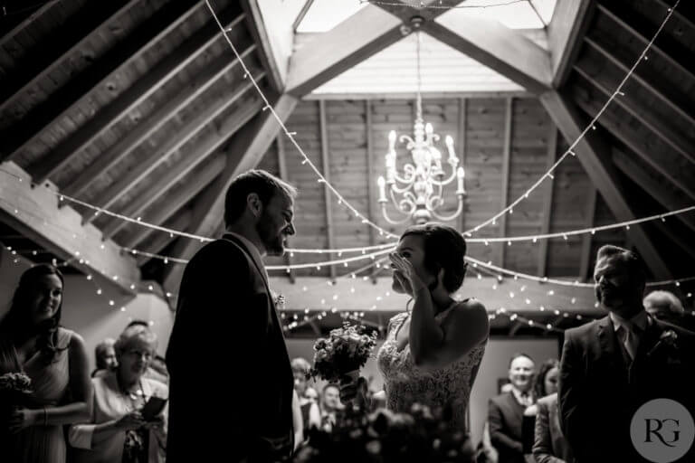 Black and white photo of bride and groom  at wedding ceremony bride wipes away tear in hotel lounge with wood panelled ceiling with string lights hanging down