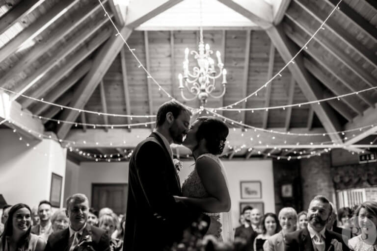 Black and white photo of bride and groom kissing at wedding ceremony in hotel lounge with wood panelled ceiling with string lights hanging down