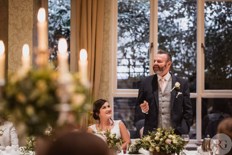 Man gives speech at wedding as bride looks up at him happily