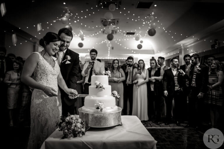 Black and white photo of bride and groom cutting wedding cake on dancefloor with wedding guests looking on and taking photos on phones