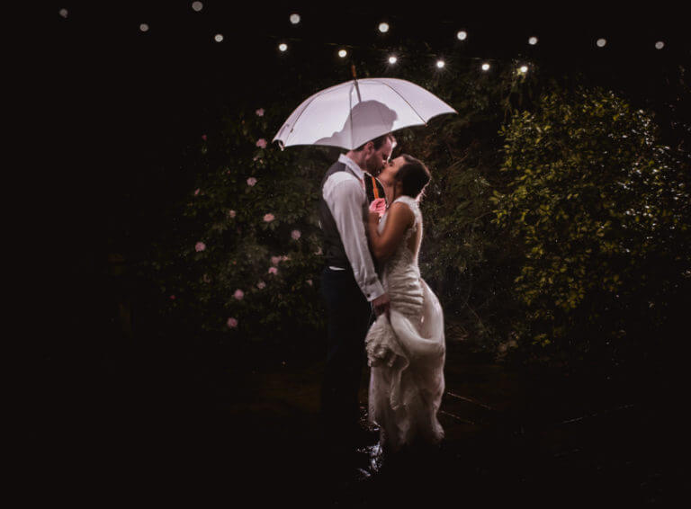 Bride and groom share a kiss under an umbrella in hotel wedding venue gardens in the evening