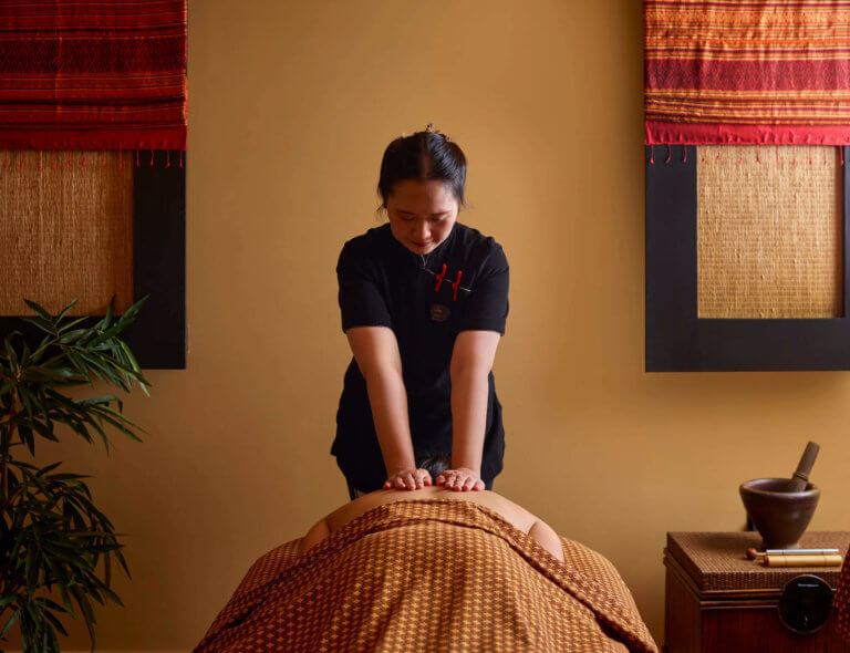A Thai massage therapist laying her hands therapeutically on her female client, who is lying down under Thai patterned sheets.