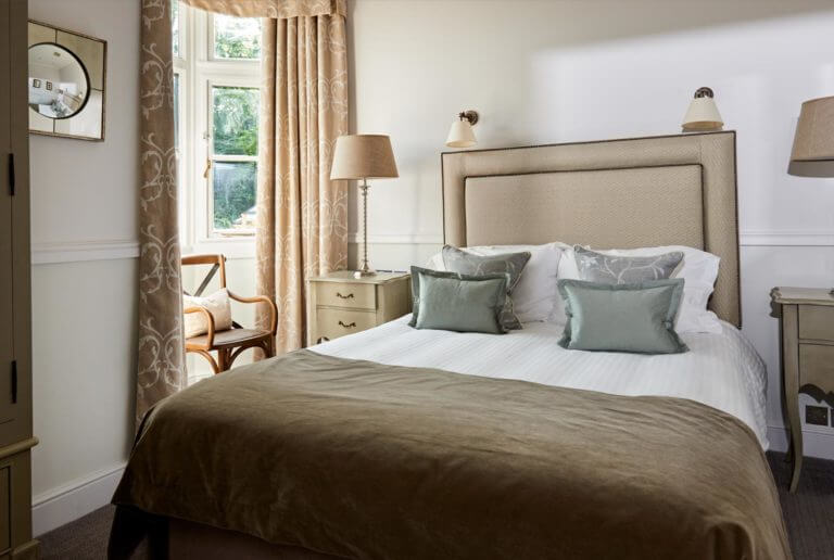 Elegant double bedroom in hotel with neutral colour theme, tall curtains with window looking out at garden