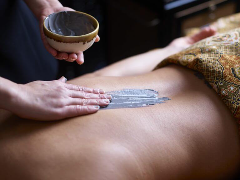 Spa therapist applies mud mask to recipient's bare back as they lie face down on massage table