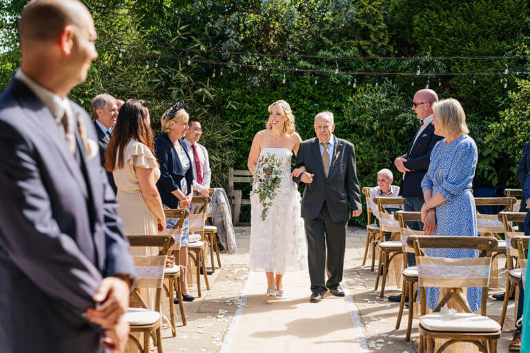 Bride is escorted up the aisle by older man as wedding guests look on happily at outdoor wedding in summer