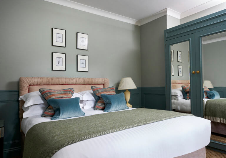 A freshly made bed with crisp white sheets, a green wool throw and blue velvet cushions. The walls are pale green with blue paneling and there is a mirrored built in wardrobe to the right