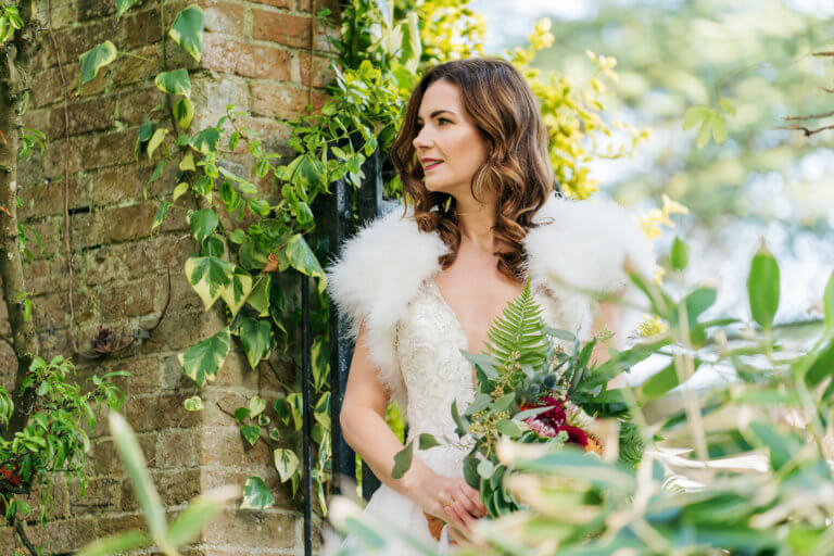 Bride in the front gardens of the hotel wearing a fur shrug and holding a red bouquet at hampshire wedding venue Careys Manor Hotel