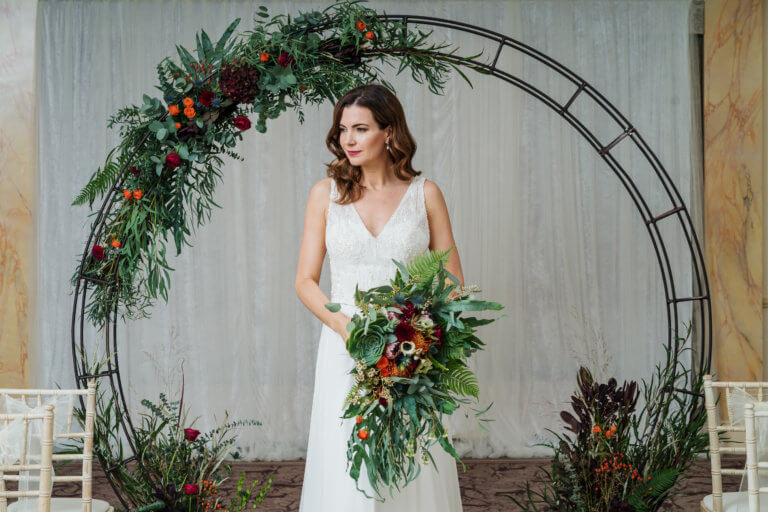 Bride stood in front of a large circular flower display holding a bouquet of red flowers at Hampshire wedding venue Careys Manor Hotel