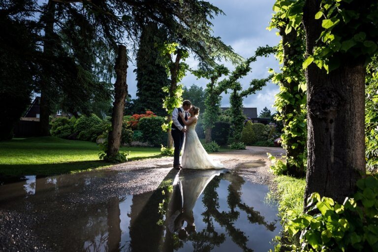 Bride & Groom at the front of the hotel in dimmed light with their reflection in a large puddle atfter the rain. Hampshire wedding venue Careys Manor Hotel