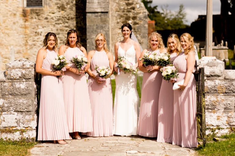 Bride Hayley surrounded by her six bridesmaids wearing floor length pink dresses outside the church