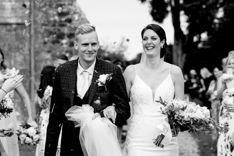 Black and white photo of bride and groom happily walking surrounded by friends and confetti