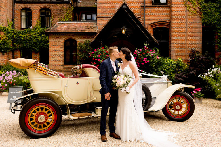 Bride and groom kiss outside hotel wedding venue entrance next to classic car