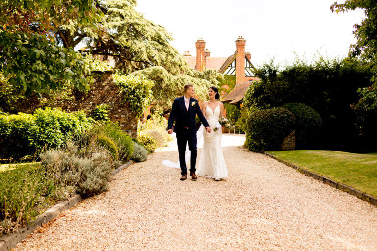 Bride and groom walk hand in hand away from hotel wedding venue through gardens 