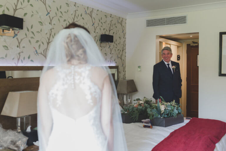 The Father of the bride gets his first glance at the bride in the bedroom to escort the bride to the ceremony at Hampshire wedding venue Careys Manor Hotel