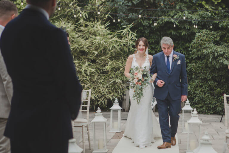 The bride is escorted by her father down the aisle of the outside wedding ceremony at New Forest Wedding venue Careys Manor Hotel