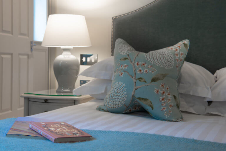 A close up of a bed in a Careys Manor suite with a duck egg blue wool throw, a floral cushion and a hard back book of Alice in Wonderland on the bed.