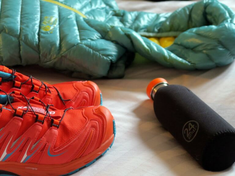 Outdoor running gear laid on floor including red trainers, black water bottle and teal padded jacket
