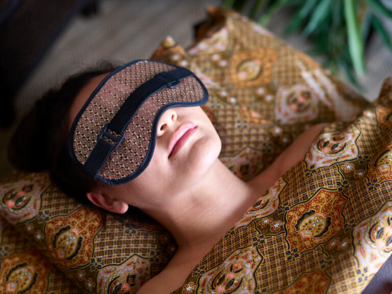 A female lying down on a therapy couch under Thai patterned sheets with an eye mask over her eyes.