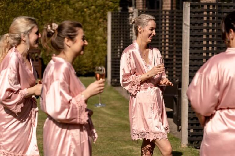 Brides maids smile and laugh whilst drinking champagne in the garden in matching robes